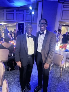 Two men in black tie attire, an older man in a black tuxedo and a younger man in a brown tuxedo jacket, stand side-by-side smiling for a photo in a dimly lit banquet hall. The room is decorated with white and blue accents, and tables are set with white linens in the background.