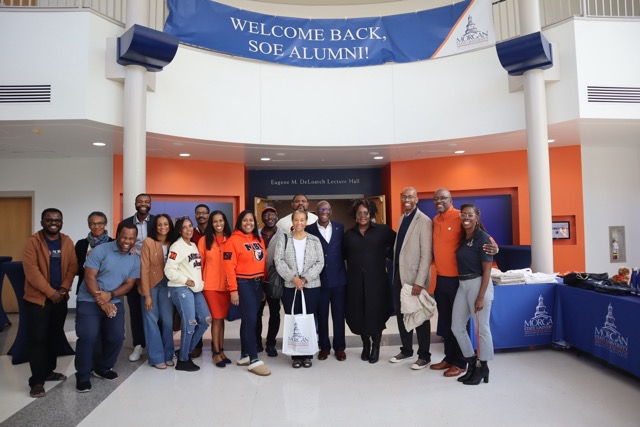 GA group of approximately 15 Morgan State University School of Engineering alumni stand smiling for a photo in a brightly lit campus building lobby beneath a banner that reads, "WELCOME BACK, SOE ALUMNI!" Many of the alumni are wearing the school colors of orange and blue.